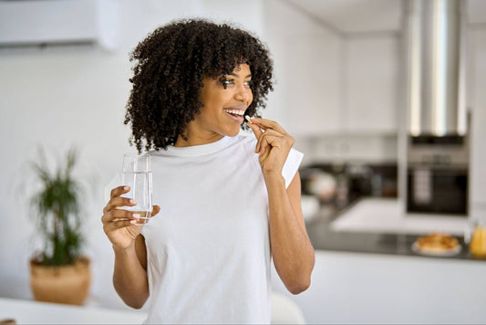 Woman in a white sleeveless shirt taking a vitamin with water at her kitchen island